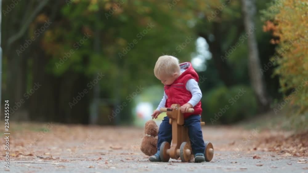 Little toddler boy with teddy bear, riding wooden dog balance bike in autumn park on a sunny warm day, children leisure activities and happiness concept Little toddler boy with teddy bear, riding wooden dog balance bike in autumn park on a sunny warm day, children leisure activities and happiness concept