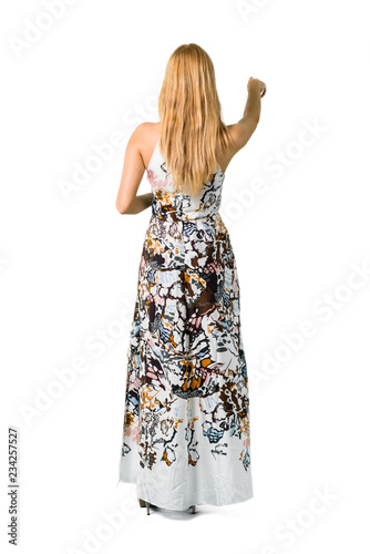 Blonde young girl in a summer dress pointing back with the index finger presenting a product from behind on isolated white backgroun