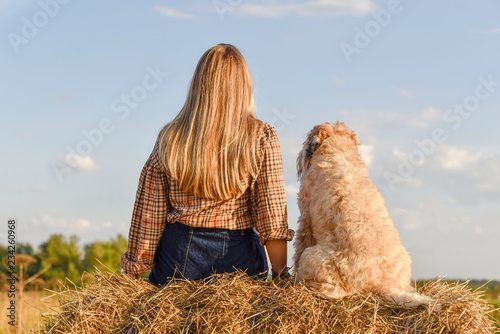 A girl with long hair sits next to a shaggy dog on a haystack with her back to the camera