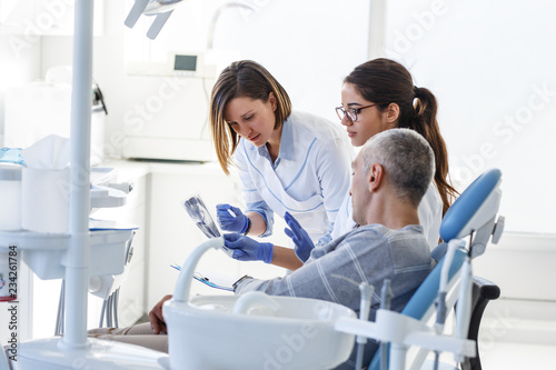 Dentist and her assistant in dental office talking with male patient and preparing for treatment.Examining x-ray image.