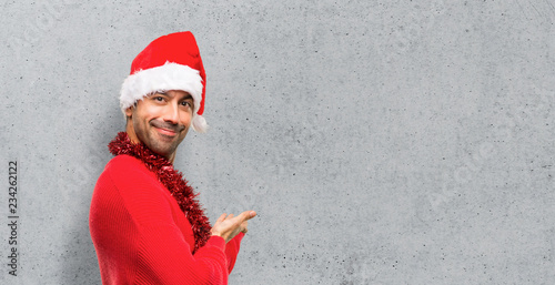Man with red clothes celebrating the Christmas holidays pointing back and presenting a product on textured background
