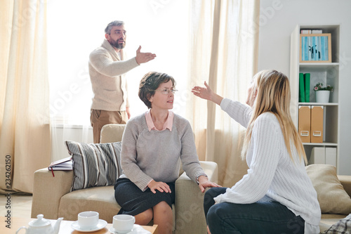 Fototapeta Naklejka Na Ścianę i Meble -  Frowning family psychologist in glasses sitting in armchair and listening to quarrel of couple while asking young lady to stop conflict, emotional couple gesturing hands and blaming each other for