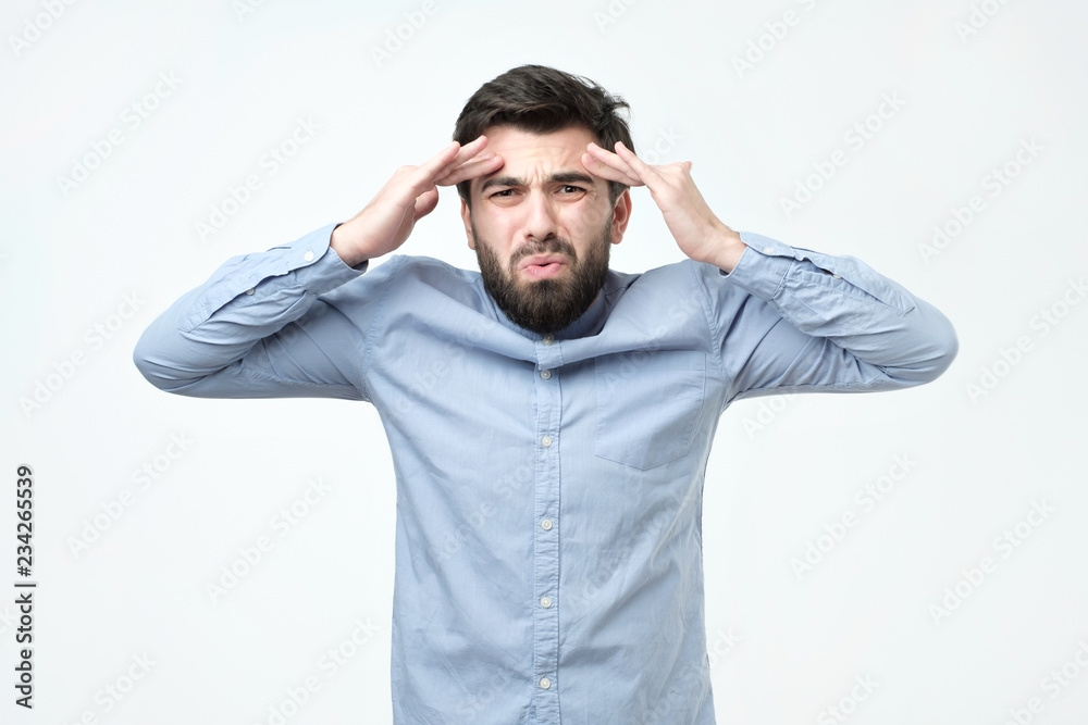 Naklejka premium Młody mężczyzna z ciemnymi włosami i brodą, ubrany w niebieską koszulę, wygląda na złego, z zaciśniętymi ustami i marszczonymi brwiami