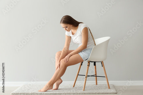 Woman suffering from pain in leg while sitting on chair near light wall