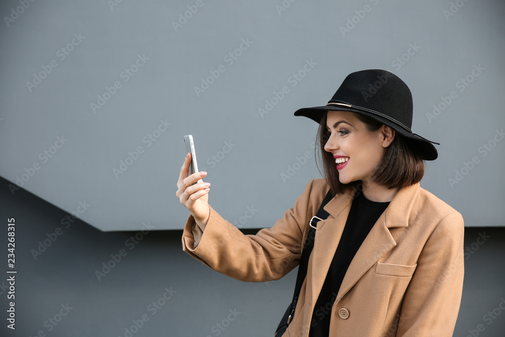 Beautiful fashionable woman with mobile phone near grey wall outdoors