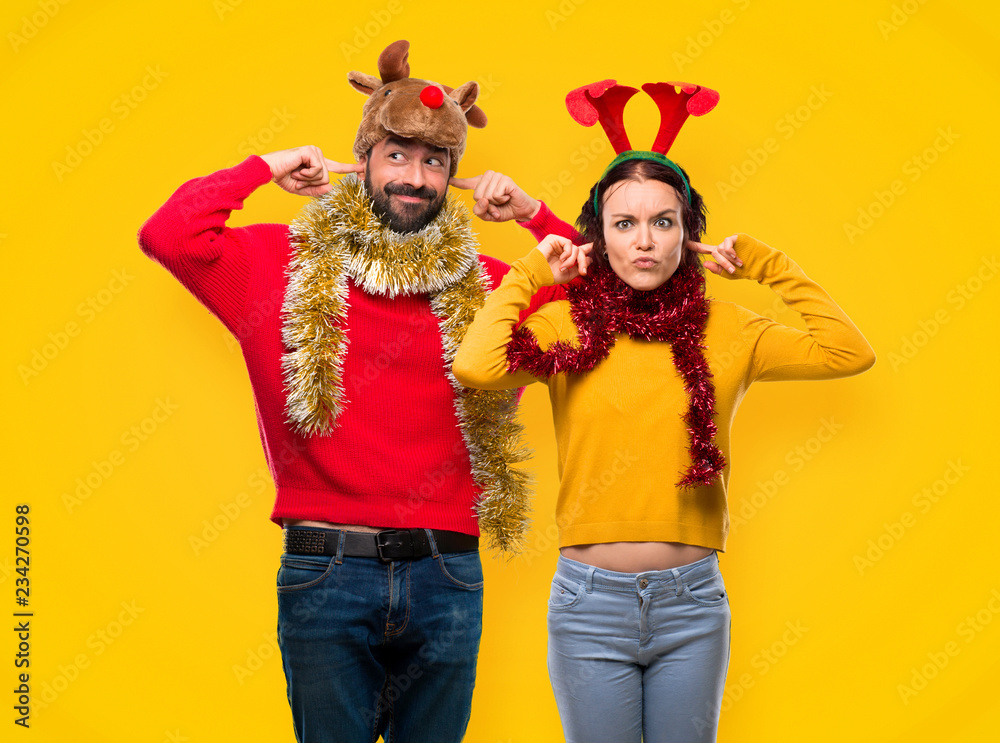 Couple dressed up for the christmas holidays covering ears with hands on yellow background Couple dressed up for the christmas holidays covering ears with hands on yellow background