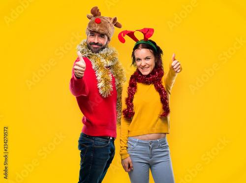 Couple dressed up for the christmas holidays giving a thumbs up gesture and smiling on yellow background