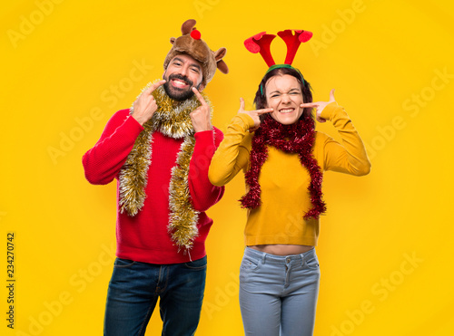 Couple dressed up for the christmas holidays smiling with a pleasant expression while pointing mouth with fingers on yellow background