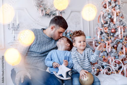 A a daddy with his two sons twins in front of the christmas tree  with candles and gifts hugging and having fun. New year's eve. Cozy holiday at the fir-tree. love, happiness and big family concept