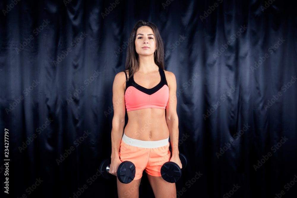 Obraz premium Fitness model in bright sports form shows exercises with dumbbells on a black background