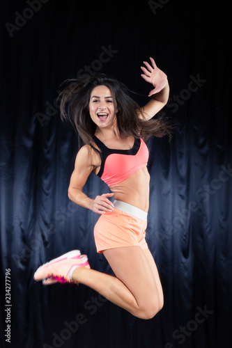 Beautiful brunette in a bright sports form with perfect body jumps on a black background. Selective focus on the face