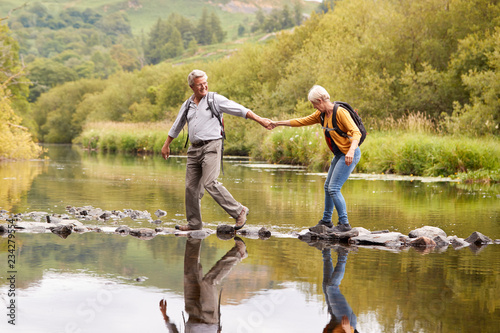 Senior Couple Crossing River Whilst Hiking In UK Lake District