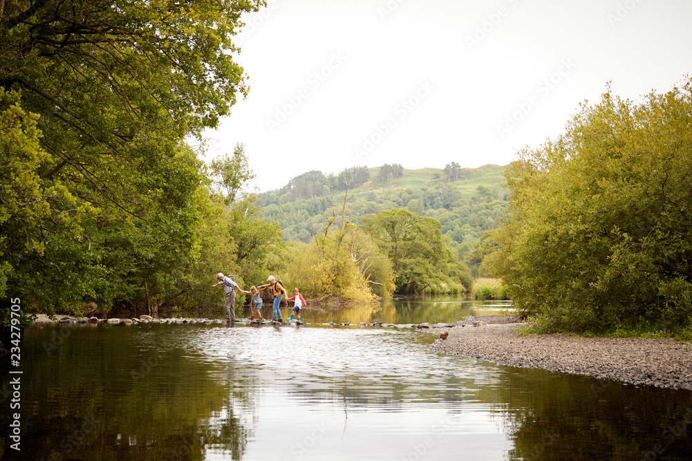 Grandparents With Grandchildren Crossing River Whilst Hiking In UK Lake District Grandparents With Grandchildren Crossing River Whilst Hiking In UK Lake District