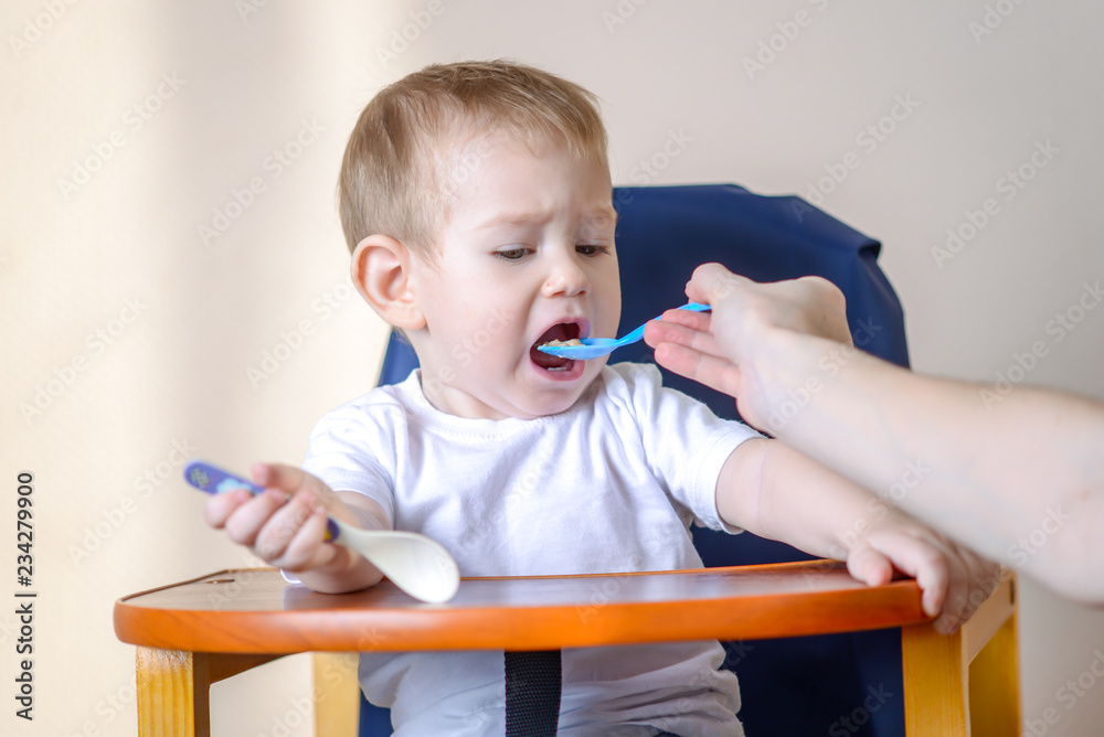 Little baby boy eats opening his mouth wide sitting on a chair in the kitchen. Mom feeds holding a spoon of porridge Little baby boy eats opening his mouth wide sitting on a chair in the kitchen. Mom feeds holding a spoon of porridge