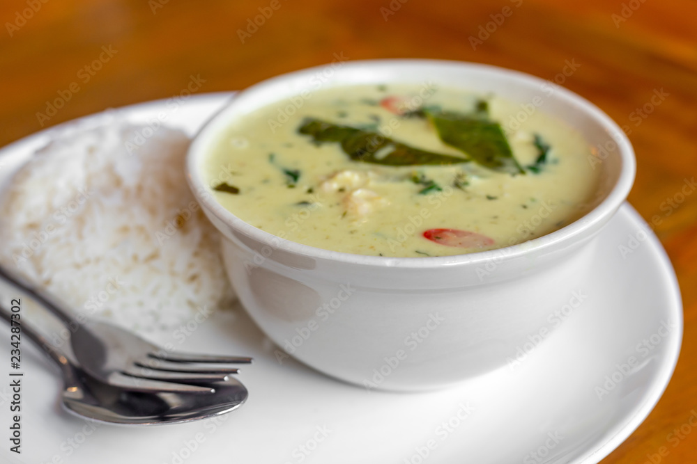 Green curry with red chili chicken and green basil leaf in the white bowl with steamed rice in the white round plate with fork and spoon on the brown wooden table. 