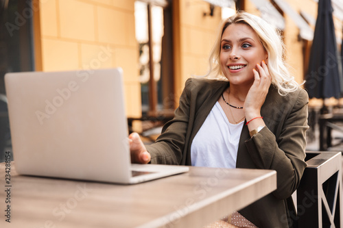 Portrait of surprised blond woman wearing jacket using silver laptop, while sitting in cafe outdoor