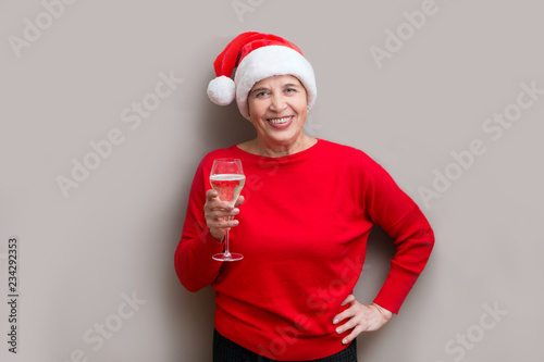Happy senior lady in Santa red hat with a glass of champagne looking at camera and smiling posing about a wall. Christmas time.