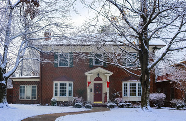 Beautiful brick house with bay windows with Christmas tree showing through and decorated pillars and sled on porch in snow framed by winter trees