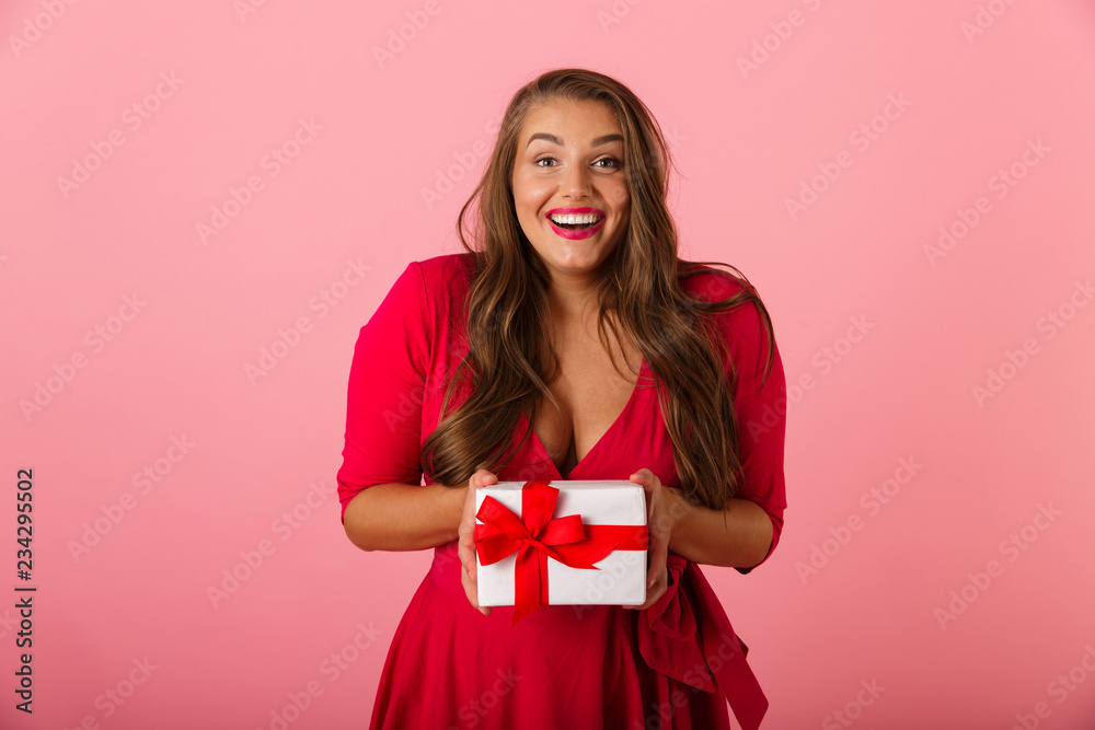 Photo of caucasian chubby woman 20s wearing red dress smiling and holding gift box, isolated over pink background Photo of caucasian chubby woman 20s wearing red dress smiling and holding gift box, isolated over pink background