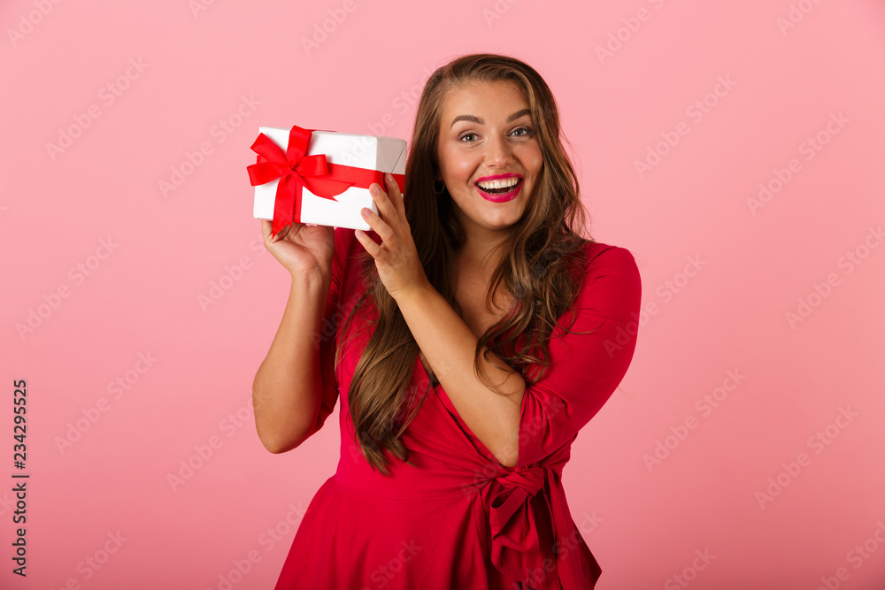 Photo of surprised overweight woman 20s wearing red dress smiling and holding gift box, isolated over pink background