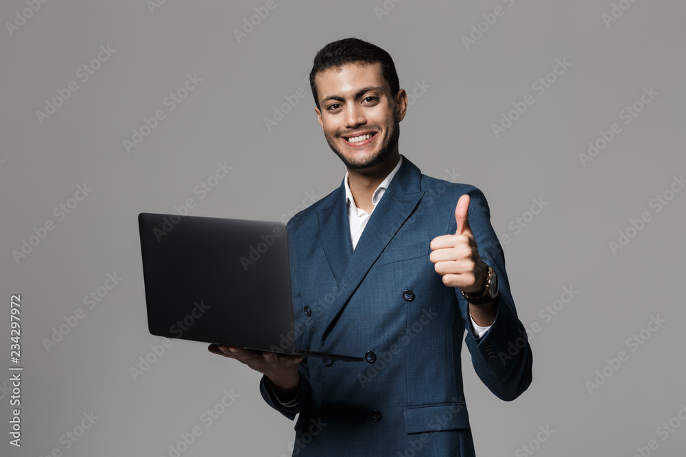 Image of brunette arabic businessman 30s in formal suit smiling and holding laptop, isolated over gray background Image of brunette arabic businessman 30s in formal suit smiling and holding laptop, isolated over gray background