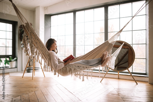 Dark-haired girl dressed in pants, sweater and warm slippers reads a book lying in a hammock in a cozy room with wooden floor and panoramic windows and a round mirror on the floor photo