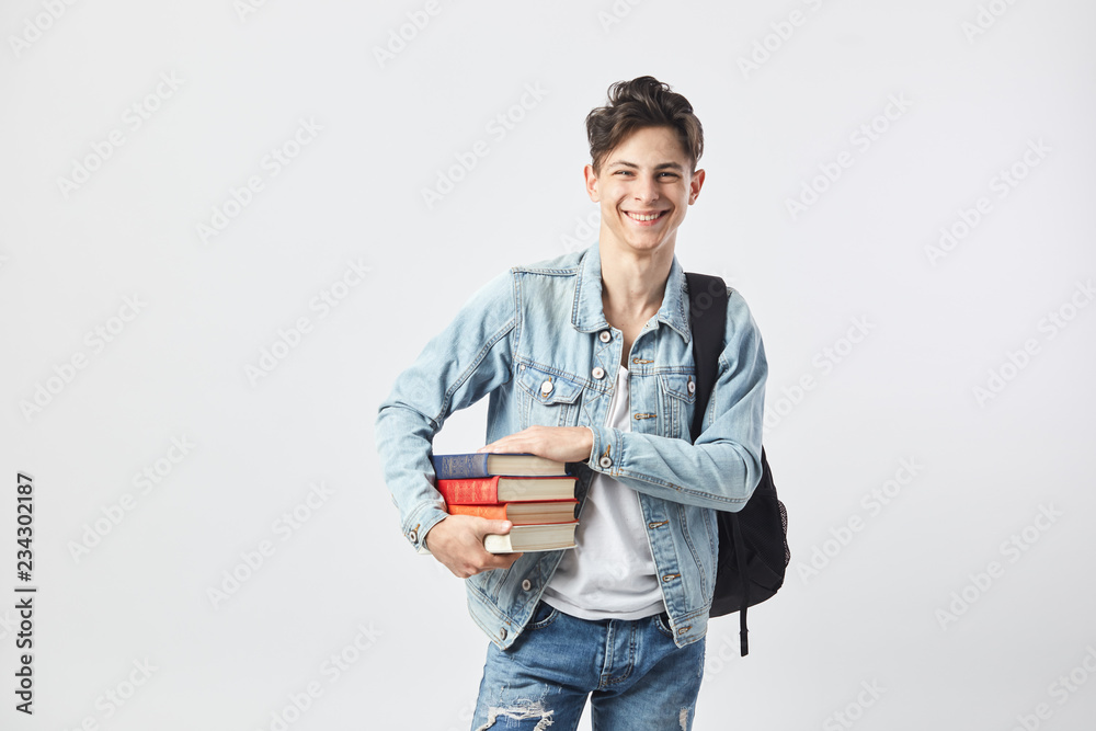 Smiling young dark-haired guy with black backpack on his shoulder dressed in a white t-shirt, jeans and a denim jacket holds books in his hands on the white background in the studio Smiling young dark-haired guy with black backpack on his shoulder dressed in a white t-shirt, jeans and a denim jacket holds books in his hands on the white background in the studio