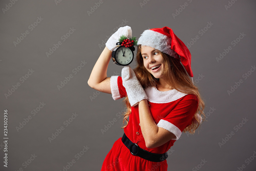 Obraz premium Beautiful mrs.Santa Claus dressed in the red robe, Santa's hat and white gloves is holding a clock that shows five to twelve. Christmas Eve