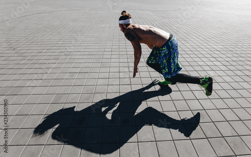 Young athletic man with a naked torso with tattoos and headband on his head dressed in the black leggings and blue shorts starting to run on paving slabs on the square on a warm sunny day