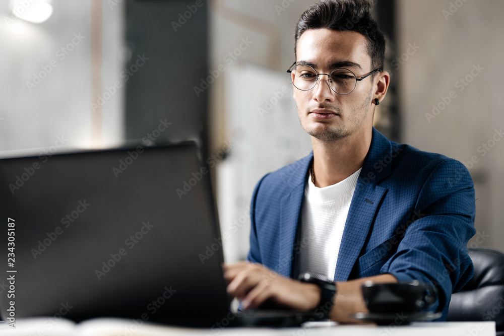 Dark-haired young architect in glasses dressed in blue checkered jacket works on the laptop in the office Dark-haired young architect in glasses dressed in blue checkered jacket works on the laptop in the office