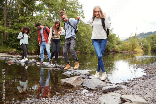 Multi ethnic group of five young adult friends hold hands walking on rocks to cross a stream during a hike #234309371