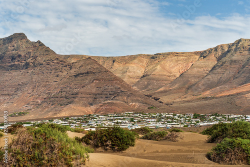 Town with white houses at the bottom of a volcanic crater, bordered by sand dunes on a sunny summer day. Caleta de Famara, coastal village in Lanzarote, Spain.