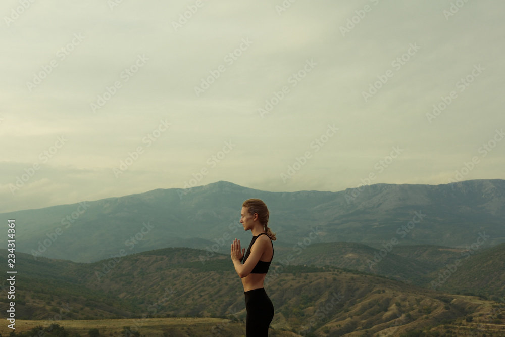 Profile portrait of slim girl dressed in black sport outfit, black top, meditating, praying, closed eyes, staying in the mountains