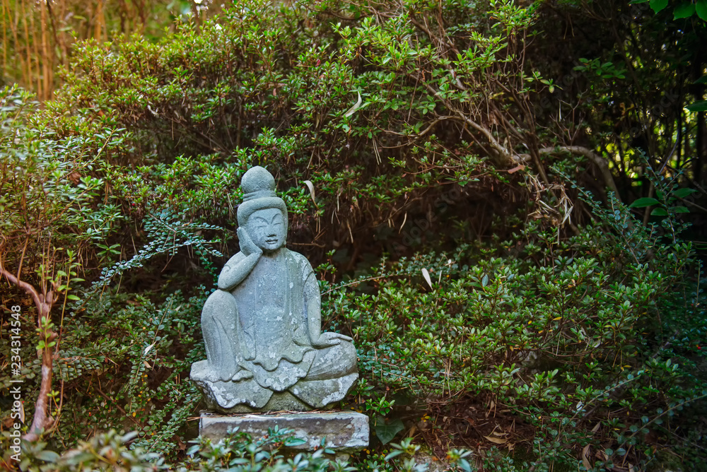 A weathered stone statue of a Buddha or Buddhist monk sitting and observing world, meditates under green trees in japanese garden. Relax and mind calm concept. Exterior,outdoor decor. Copy space.