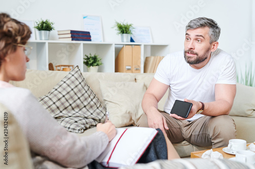 Furious emotional handsome bearded man sitting on sofa with cushions and showing video of wives adultery while sharing his sufferings with psychologist