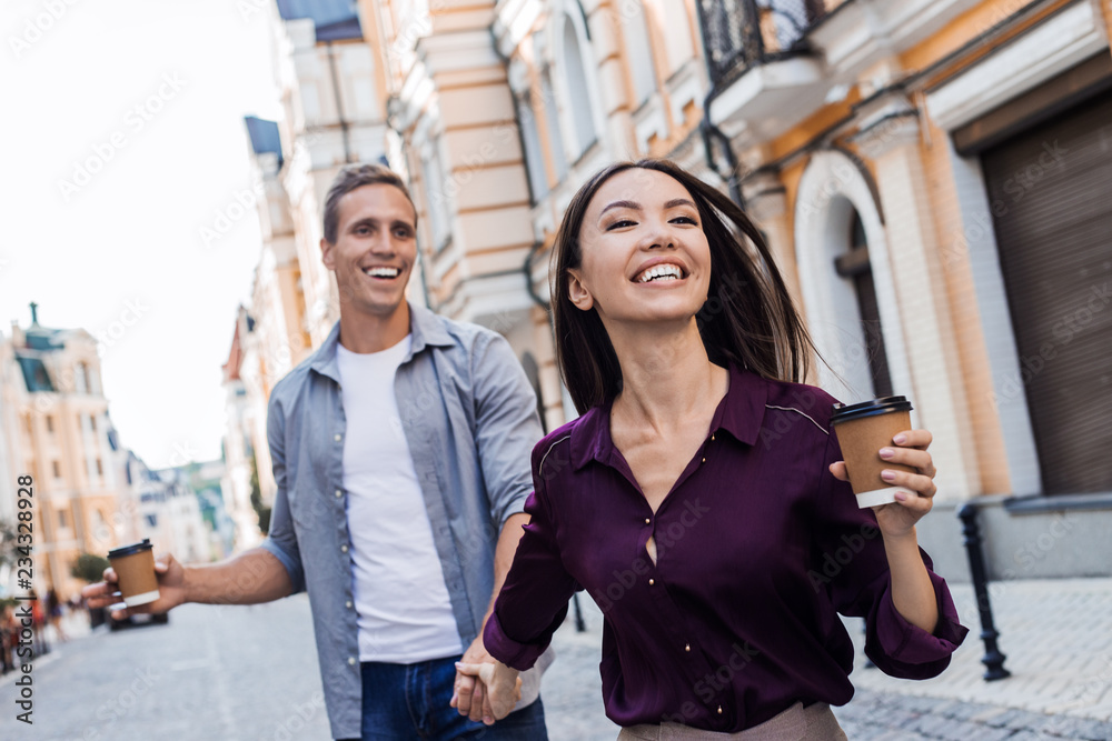 Beautiful woman holds boyfriend by the hand and goes with him along the street of the old town on the background of the sunset. Happy man follows his girlfriend. Follow me concept, female perseverance