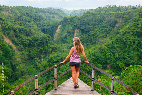 Family vacation lifestyle. Young woman stand on edge of overhanging bridge on high cliff. Happy girl looking at stunning tropical jungle view. Tukad Melangit is popular travel destination in Bali. photo