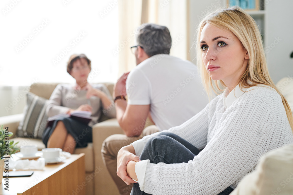 Sad thoughtful attractive young wife with blond hair sitting on sofa and embracing her knees while her husband talking to psychiatrist at therapy session