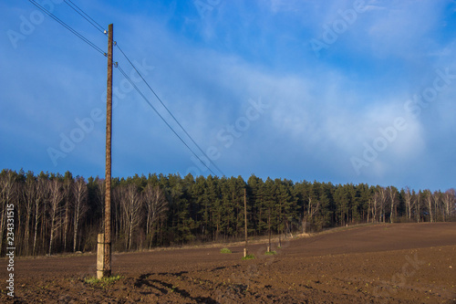 a line of high-voltage pillars through a plowed agricultural field, a forest on the horizon and a blue sky
