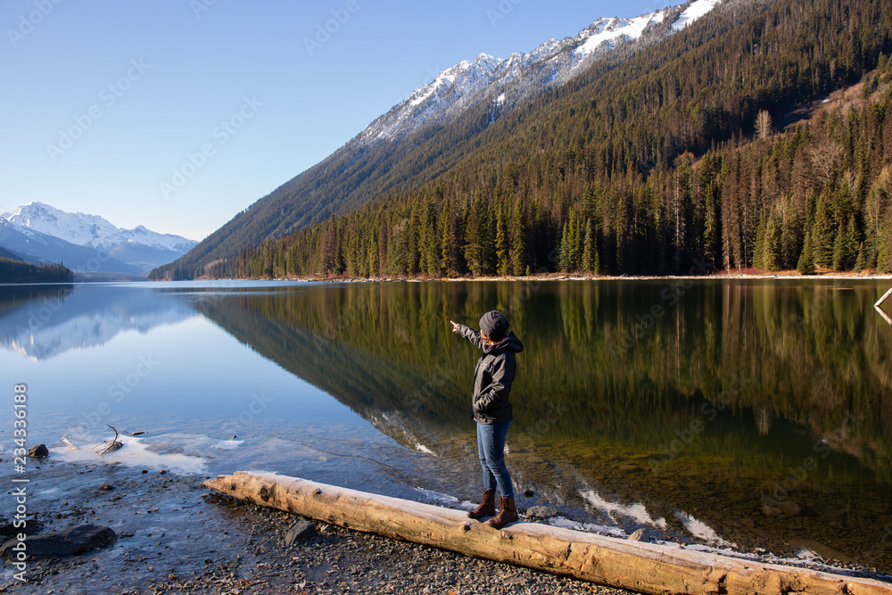 British Columbia Canada, Duffy Lake with smooth water reflection and mountains in backgrounds and woman standing on log pointing 