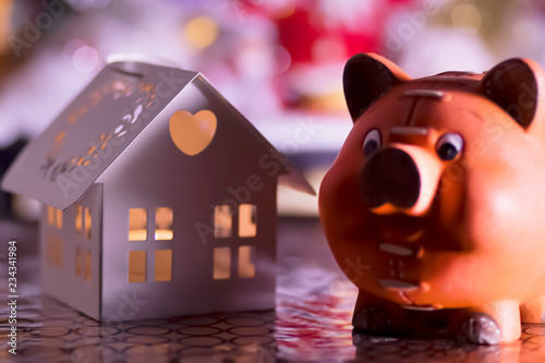 Pig figurine and a candlestick in the form of a white house on the background of Christmas decorations.