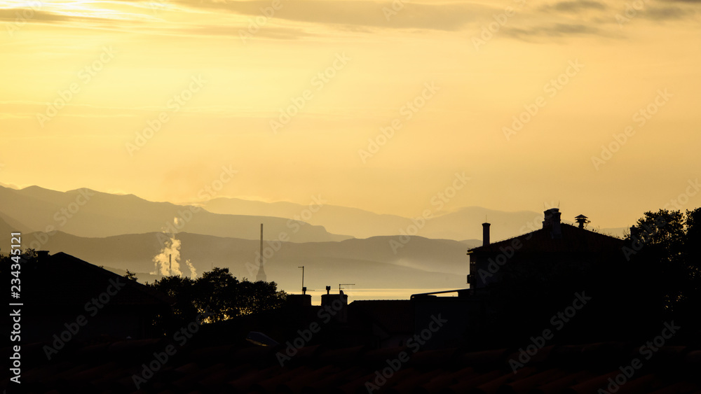Early morning in an industrial city, house silhouettes in foreground, and layered mountains covered with clouds, smoke and smog in the background; black and orange landscape