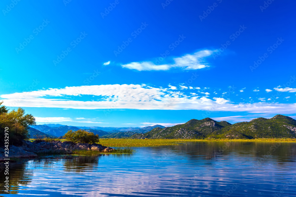 Obraz premium The beautiful smooth surface of the blue lake reflects the blue sky with white clouds, hilly terrain with trees among the stone slopes on the shore. Skadar Lake, Podgorica region, Montenegro.