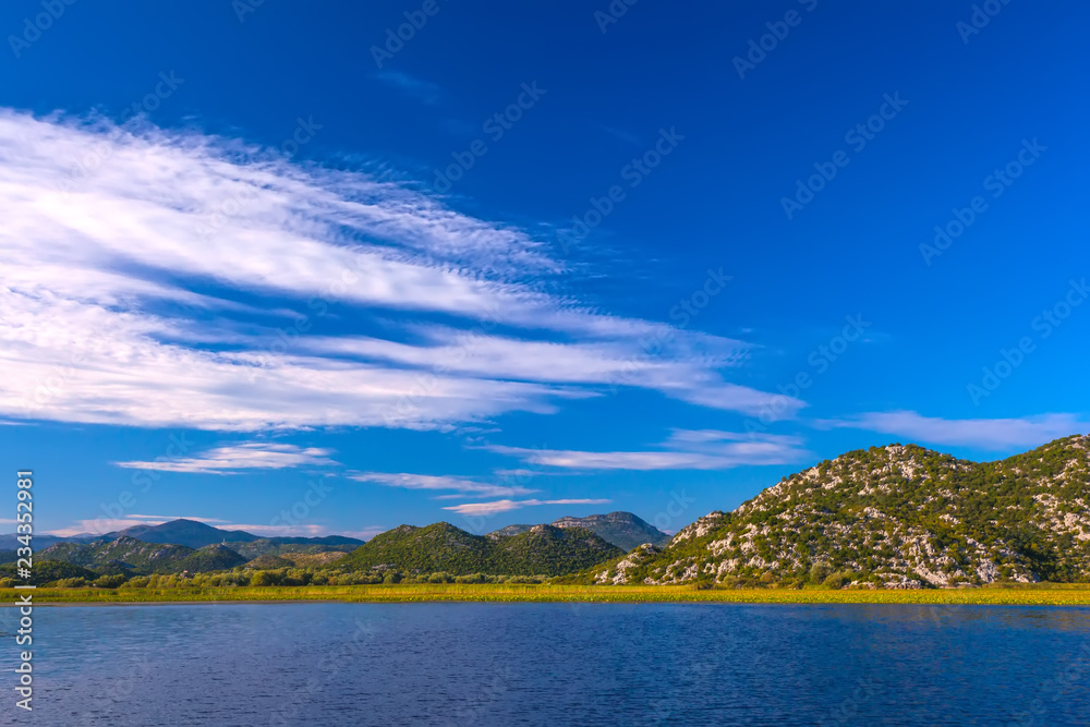 The beautiful smooth surface of the blue lake reflects the blue sky with white clouds, hilly terrain with trees among the stone slopes on the shore. Skadar Lake, Podgorica region, Montenegro.