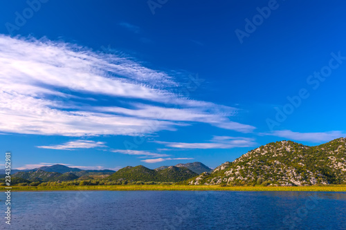 The beautiful smooth surface of the blue lake reflects the blue sky with white clouds, hilly terrain with trees among the stone slopes on the shore. Skadar Lake, Podgorica region, Montenegro. © Valery Bocman