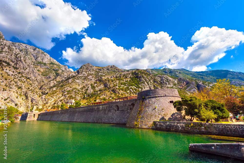 Large historical medieval stone wall in front of a moat with green water on the backdrop of the picturesque mountains on a sunny day. Kotor Venetian fortifications, Kampana Tower Old Town, Montenegro.