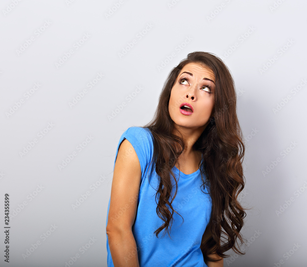 Beautiful young makeup surrising woman with curly hairstyle looking up with open mouth on blue copy empty space background Beautiful young makeup surrising woman with curly hairstyle looking up with open mouth on blue copy empty space background