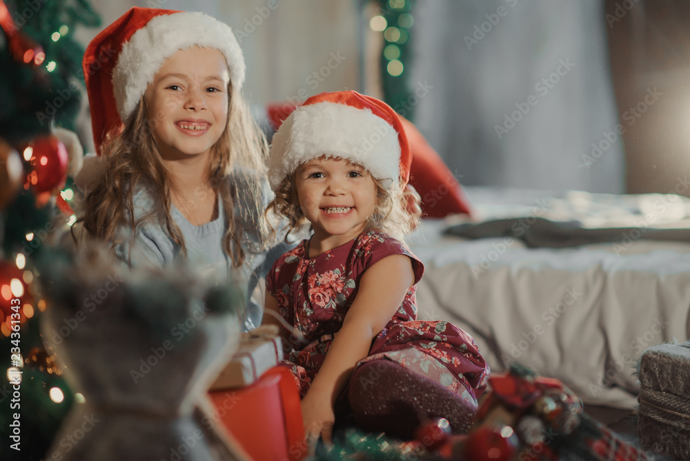 Merry Christmas and Happy Holiday! Two cute little children girls sisters open magic gift box on Christmas. Cozy room with New year decoration and Christmas tree Merry Christmas and Happy Holiday! Two cute little children girls sisters open magic gift box on Christmas. Cozy room with New year decoration and Christmas tree