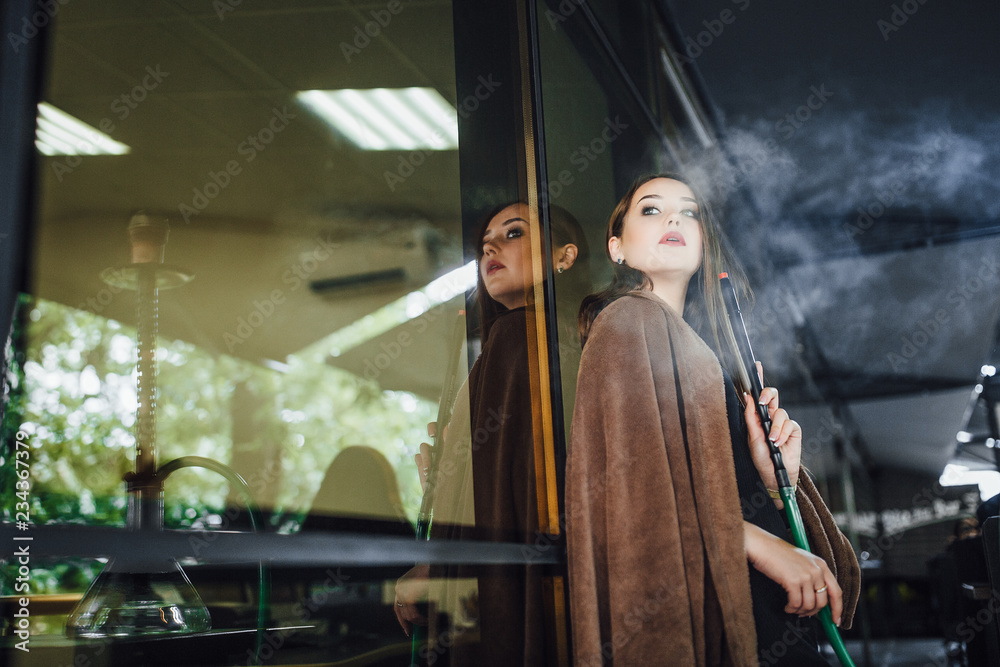 The young beautiful girl is covered with a blanket, stands and smokes hookah on the summer terrace of the modern restaurant. Dressed in a little black dress.