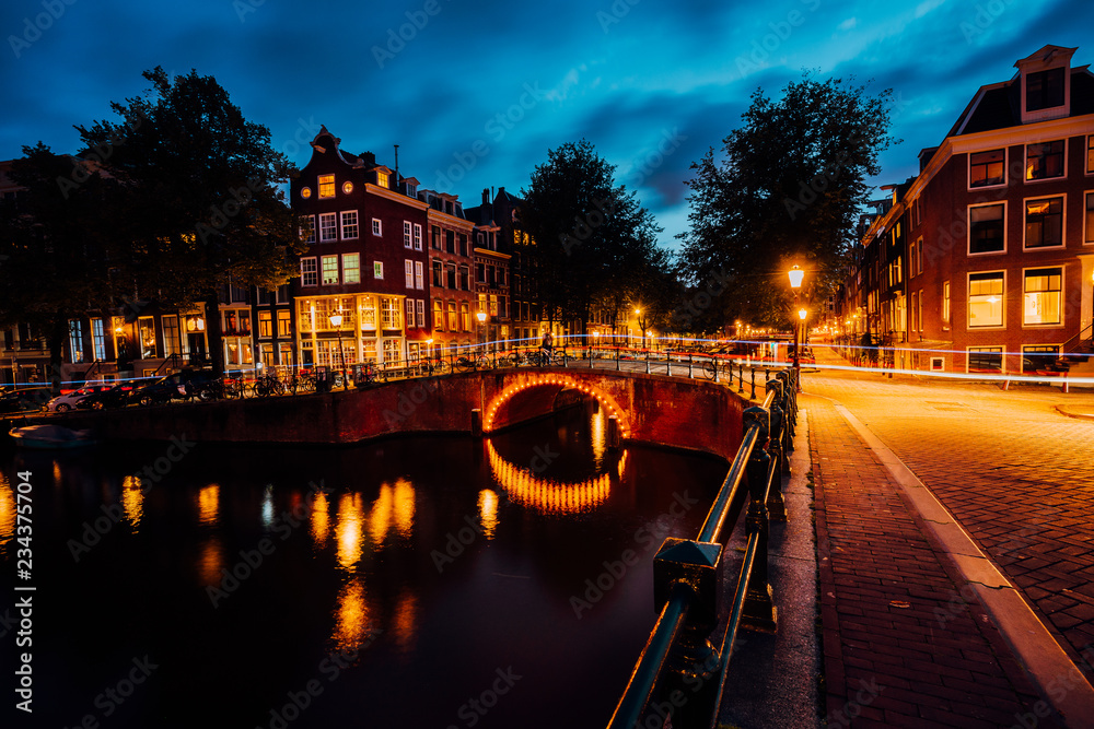 Amazing night in Amsterdam. Illuminated canal and bridge with typical dutch houses and bicycles, Holland, Netherlands. Lights trails from the bicycles. Long exposure